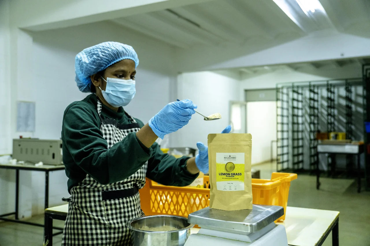 Mitarbeiterin in Schutzkleidung wiegt Bio-Zitronengras-Pulver mit Löffel in fertige MrCOLOMBO Papiertüte auf Edelstahl-Waage in Sri Lanka